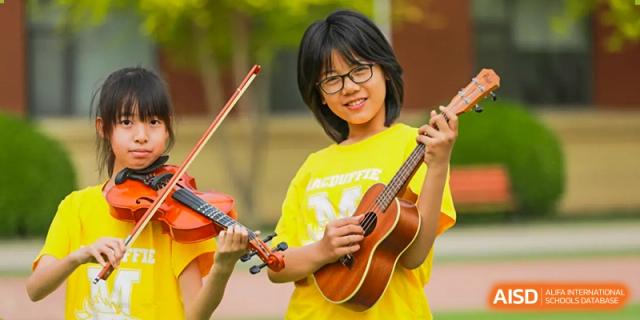 A female student in a yellow shirt is smiling while holding a ukulele and a violin at The MacDuffie School Tianjin.