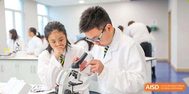 A male Wellington College International Hangzhou student wearing a lab coat and glasses, intensely focused on using a microscope during a science class.