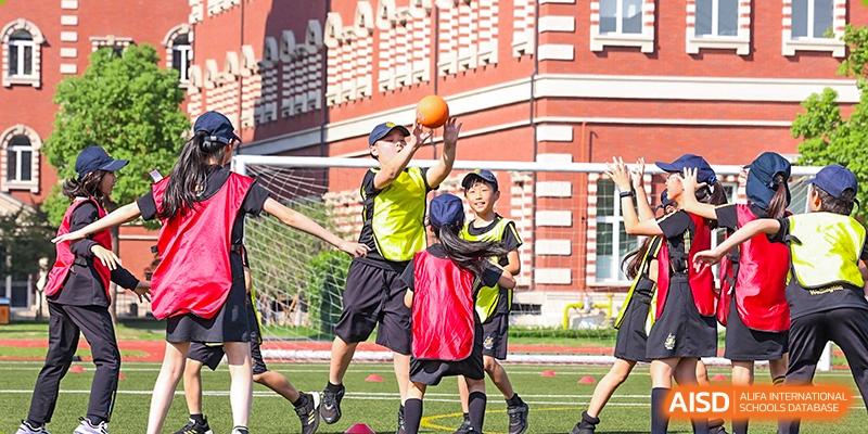 Students from Wellington College International Hangzhou playing sports, with one student attempting to catch or shoot an orange ball near a soccer goal on a field with a red-brick building in the background.