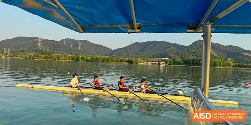 Wider shot of two students from Wellington College International Hangzhou rowing a boat on a calm lake, with green hills and buildings visible in the distance under a bright sky.