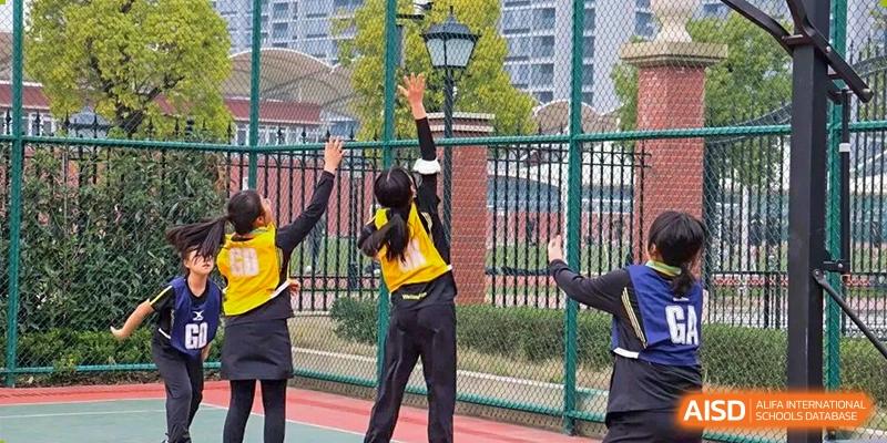 Student from Wellington College International Hangzhou reaching to hit or block a ball over a net in an outdoor court setting, demonstrating a volleyball or netball skill.