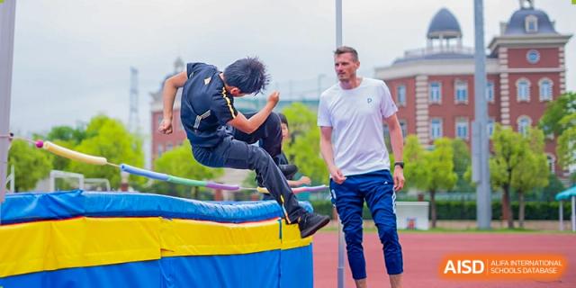 A student performing a high jump or hurdle on an athletics track, with a coach or instructor watching nearby. Wellington College International Hangzhou