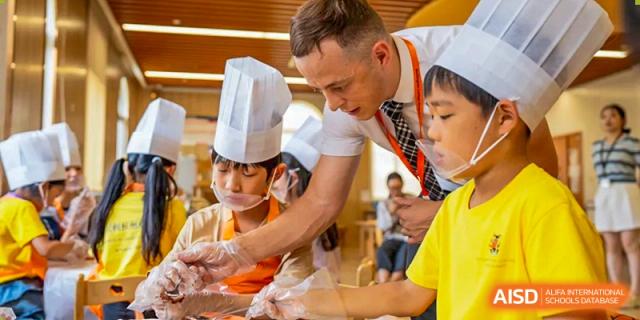 A teacher assisting two young Wellington College International Hangzhou students during a cooking or food class; the students are wearing chef hats, masks, and gloves.