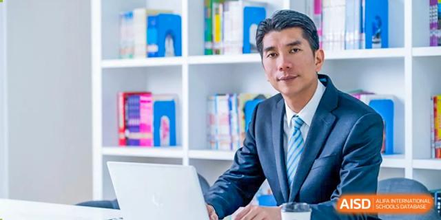 A formal portrait of a male teacher or administrator from Zhejiang Wenzhou High School International Division wearing a suit and tie, seated in a classroom with bookshelves.