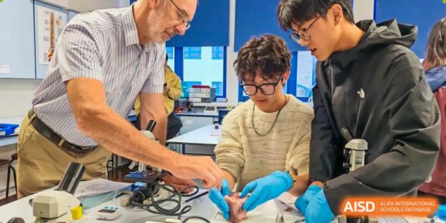 A Zhejiang Wenzhou High School International Division student wearing safety gloves and goggles, focused on a hands-on procedure with a male teacher supervising closely.