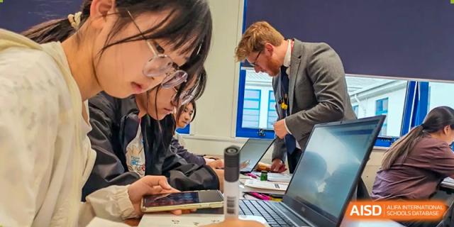 A male teacher assisting two Zhejiang Wenzhou High School International Division students with work on a laptop and checking a microscope in a classroom setting.