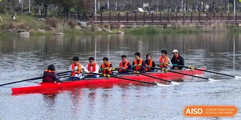 Students from Zhejiang Wenzhou High School International Division in orange life vests rowing a red boat on a calm lake as part of a sports team or club activity.