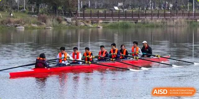Students from Zhejiang Wenzhou High School International Division in orange life vests rowing a red boat on a calm lake as part of a sports team or club activity.