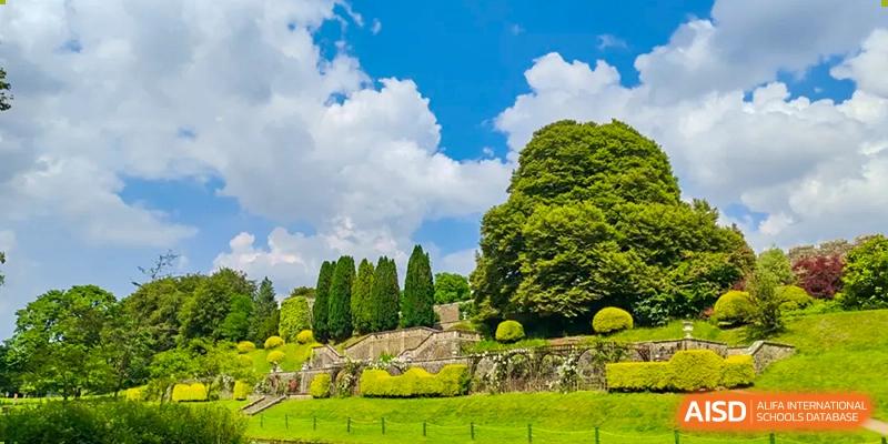 A beautiful, sunny landscape photo showing well-maintained, lush green gardens and trees under a bright blue sky, possibly representing the grounds of Zhejiang Wenzhou High School International Division.