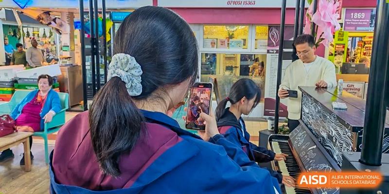 A student from Zhejiang Wenzhou High School International Division wearing a blue uniform, holding a mobile phone up to take a picture or video in a bright, indoor public area.