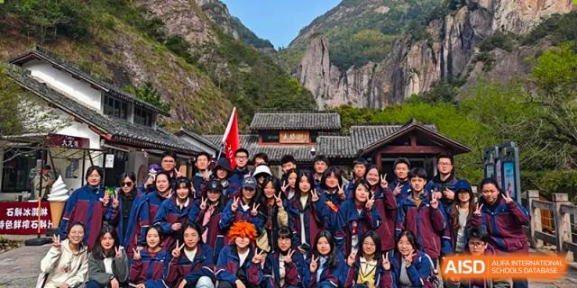 Students from Zhejiang Wenzhou High School International Division posing for a group photo in front of a traditional Chinese building nestled in a mountainous outdoor setting during a trip.