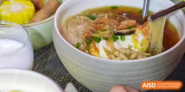 A close-up shot of a large bowl of traditional Asian noodles (vermicelli) with vegetables, egg, and broth, served in the dining hall of Zhejiang Wenzhou High School International Division.