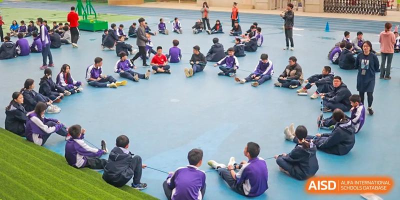 A group of students in purple jackets sits on the ground while watching an outdoor activity. Guangzhou International Primary/Middle School Huangpu