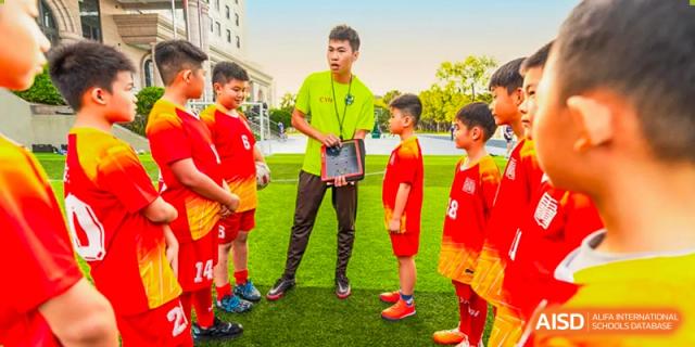 A teacher in a green shirt and a visor stands with students in red jerseys on a soccer field. Guangzhou International Primary/Middle School Huangpu