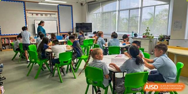 A bright classroom at Zhuhai Qiao International School with several students in uniform seated at desks with bright green chairs, looking toward a screen in the front.