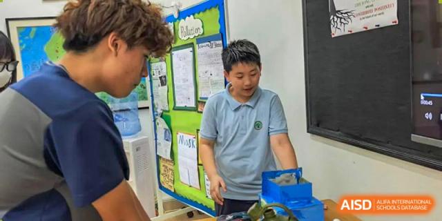 A male student at Zhuhai Qiao International School presenting a project or demonstration using a poster board and a blue box.