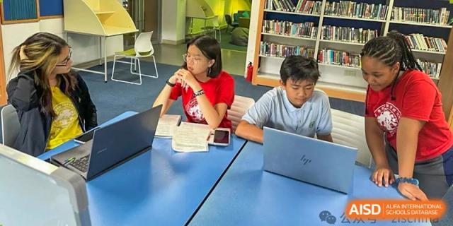 Students at Zhuhai Qiao International School working collaboratively at a blue table, using a laptop and notebooks.