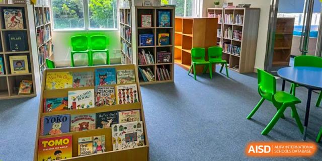 A well-stocked library or book corner at Zhuhai Qiao International School, featuring wooden bookshelves filled with books and two bright green children's chairs.