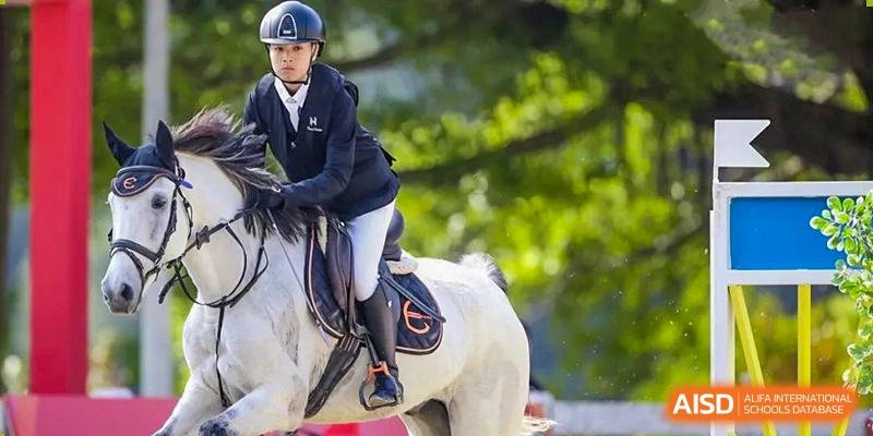 A student from Zhuhai Qiao International School demonstrating equestrian skills while riding a small white horse, wearing safety gear.