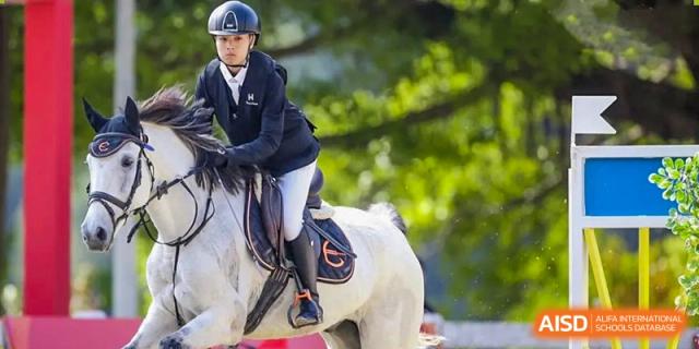 A student from Zhuhai Qiao International School demonstrating equestrian skills while riding a small white horse, wearing safety gear.