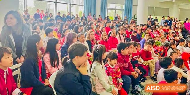 A large gathering of the Zhuhai Qiao International School community, including many students in red, attending a school assembly or performance.