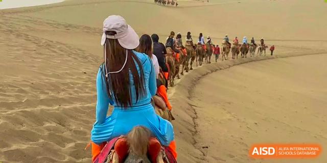 A student from Zhuhai Qiao International School riding a camel in a desert or beach setting during an educational field trip.