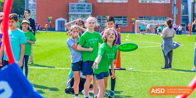 A group of younger Concordia International School Shanghai students, wearing green shirts, playing an outdoor game involving throwing a frisbee.