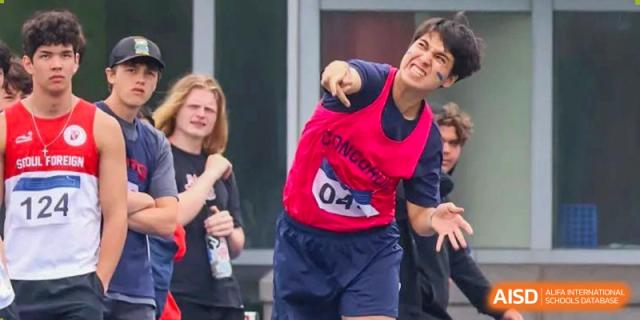 A male student from Concordia International School Shanghai, wearing a red sports vest, throwing a ball or object during an athletic event.