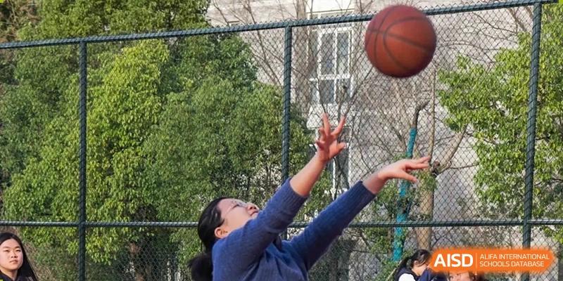 A female student reaching up to catch a basketball on an outdoor court fenced with a chain-link fence at the Hefei No.1 High School.