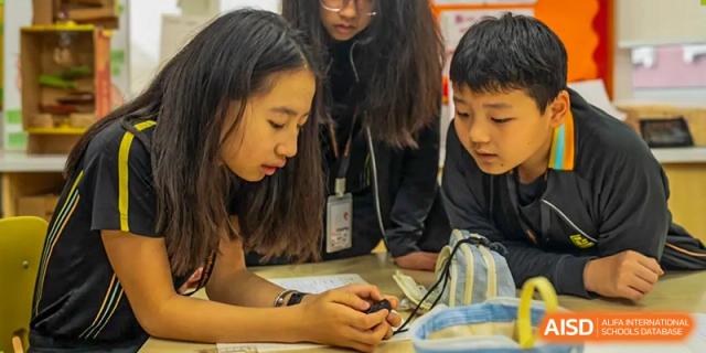 A group of students leaning over a table and examining a small electronic device or experimental apparatus during class at Hiba Academy Hangzhou.