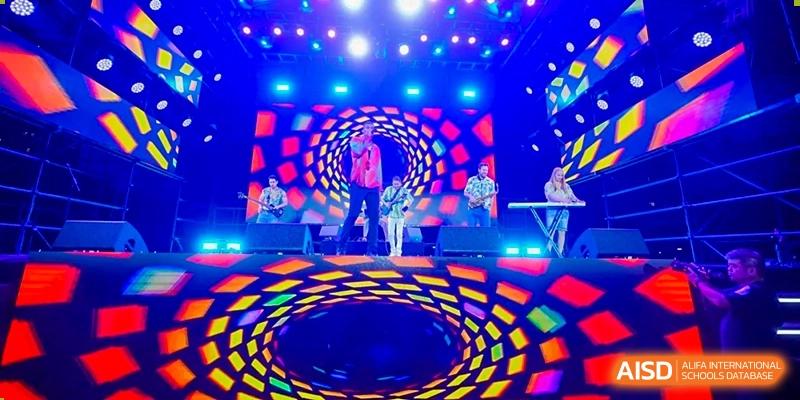 A brightly lit stage with a large LED screen showing a swirling pattern and performers during a vibrant school event at Hiba Academy Hangzhou.