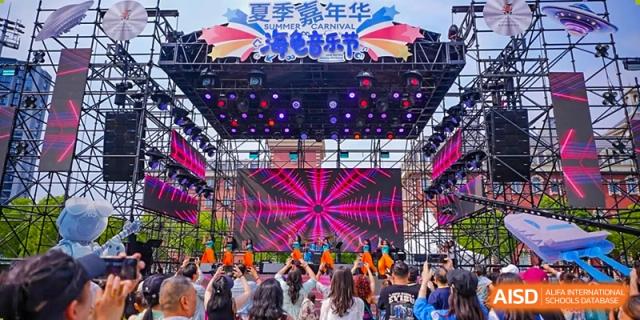 A large crowd of people attending an outdoor summer carnival event under a stage sign that reads in Chinese "夏季嘉年华" (Summer Carnival) at Hiba Academy Hangzhou.
