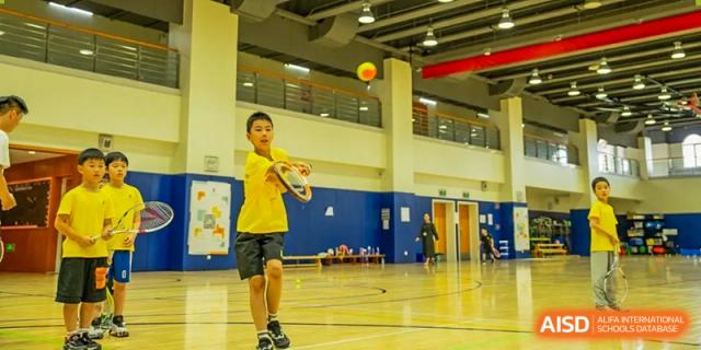 A male student in a yellow shirt playing badminton on an indoor gymnasium court at Hiba Academy Hangzhou.