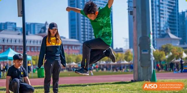 A male student in a green shirt performing a long jump outdoors with city buildings in the background at Hiba Academy Hangzhou.