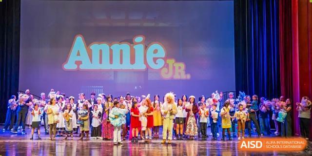 A large cast of students in bright costumes posing on stage in front of a screen displaying the title Annie Jr. at Hiba Academy Hangzhou.