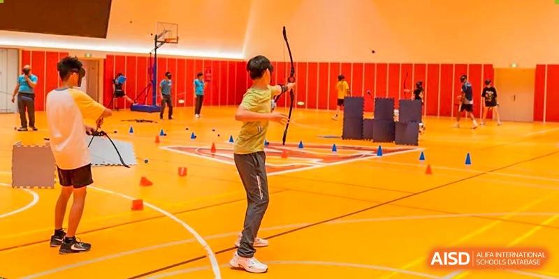 A student practicing archery indoors, aiming at a target in a large, open gym at Huitong School & Studios.