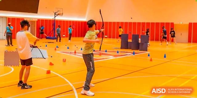 A student practicing archery indoors, aiming at a target in a large, open gym at Huitong School & Studios.