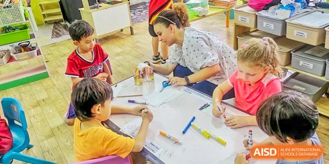 A female teacher at QSI International School of Zhuhai helps students with a hands-on art or writing project at a large table.