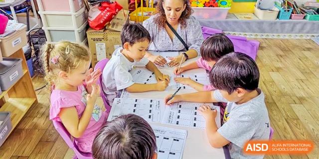 A female teacher at QSI International School of Zhuhai helps two young students with a tracing or writing activity at a table.