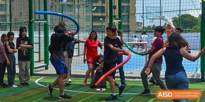 Students and a teacher from QSI International School of Zhuhai smiling and participating in a team-building activity on an outdoor sports pitch.