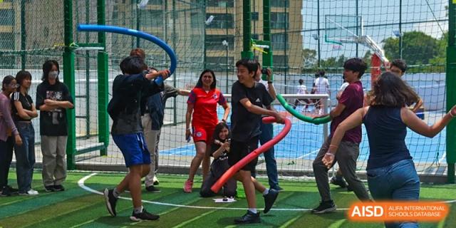 Students and a teacher from QSI International School of Zhuhai smiling and participating in a team-building activity on an outdoor sports pitch.