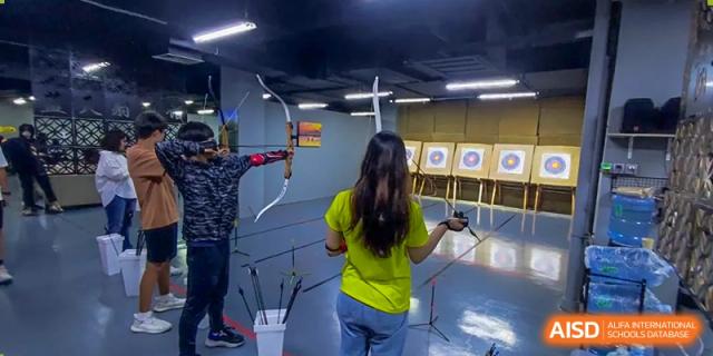 A female student at QSI International School of Zhuhai practices archery in an indoor range, aiming at a target.