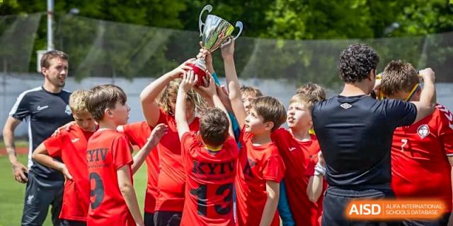 Young male students from QSI International School of Zhuhai in red jerseys cheer as they hold a large trophy high above their heads on the sports field.