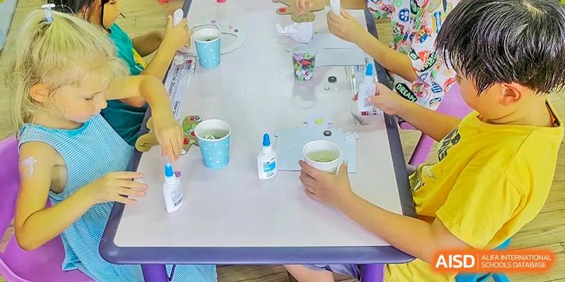 Students at QSI International School of Zhuhai participate in a hands-on art or craft activity, working with small cups and glue at a table.