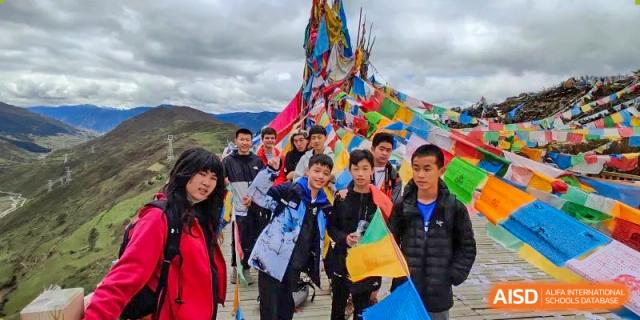 A group of students from QSI International School of Zhuhai stands together outdoors, holding colorful prayer flags during a cultural or outdoor education trip.