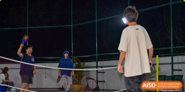 Two students at QSI International School of Zhuhai play tennis on an outdoor court at night under floodlights.