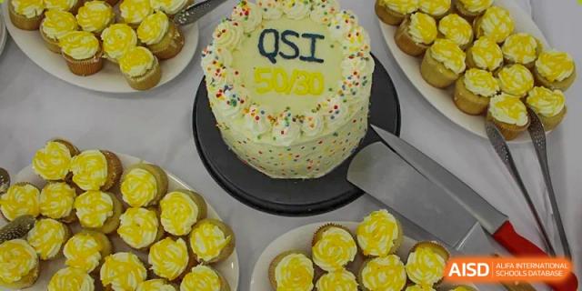A large white celebration cake decorated with colorful sprinkles and mini cupcakes, possibly for a school event at QSI International School of Zhuhai.