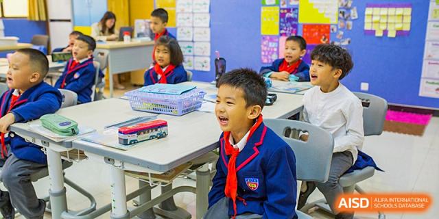 A young male student in a blue school uniform and red scarf is seen with a wide-open, excited expression while sitting at a desk in class at Wuhan Aoxin Elite School.