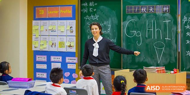 A female teacher smiling and speaking to a group of young students in a classroom with a chalkboard behind her at Wuhan Aoxin Elite School.