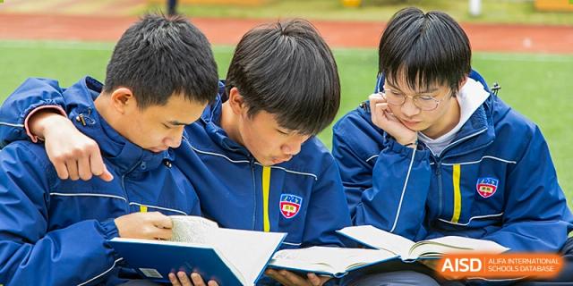 A male student in a blue school uniform with a yellow stripe intently reading a book outdoors or in a bright common area at Wuhan Aoxin Elite School.